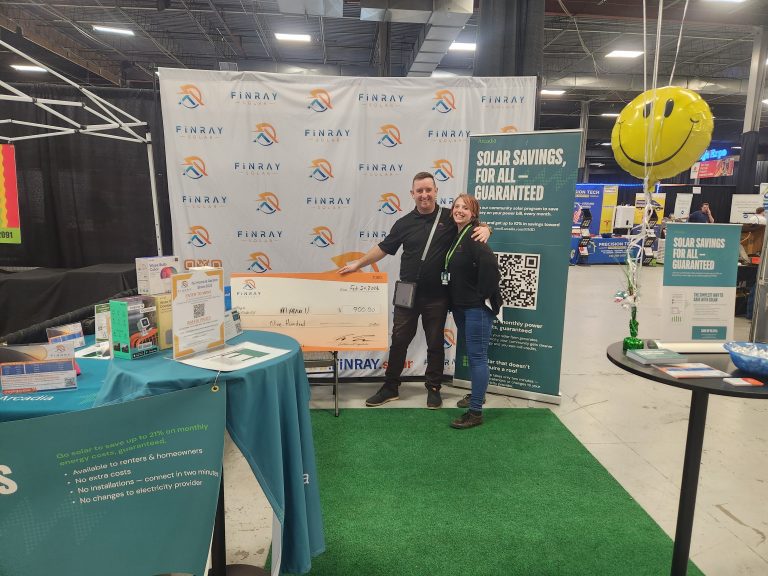 Two of our team members standing in front of our Finray Solar event banners, holding a large novelty check showing a customer's estimated electricity savings over 5 years, written in marker. In front,  tables with documents and pamphlets related to the service. Large green faux-grass rug lays on the conference floor.